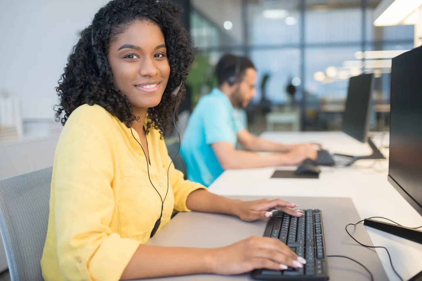 A headset-wearing agent at a keyboard in an open-plan office, with a colleague taking a call behind.